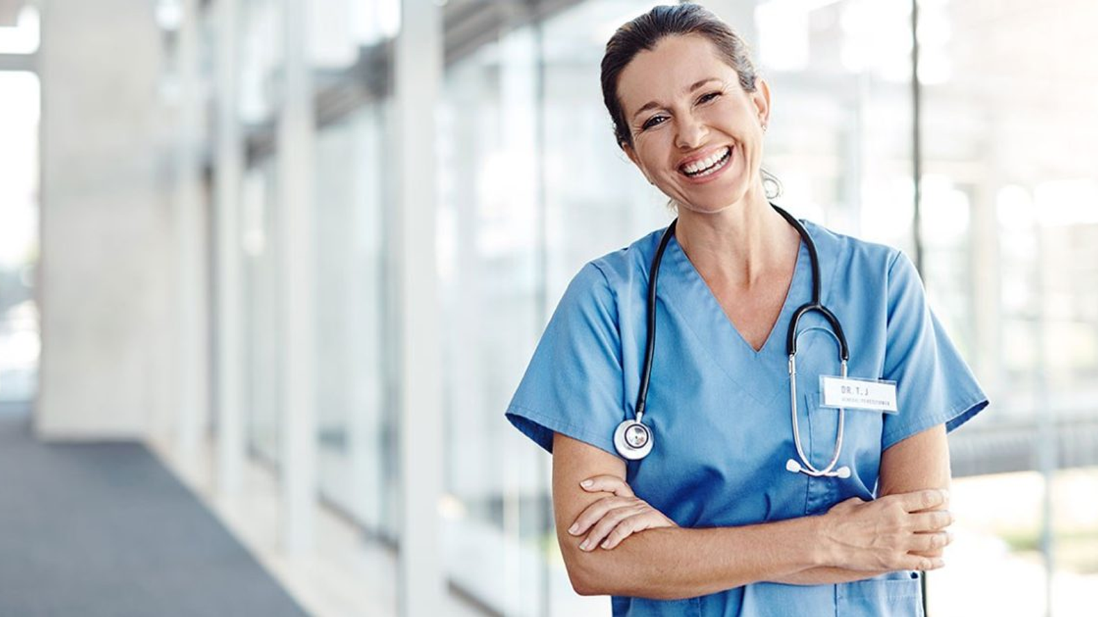 A smiling healthcare professional in blue scrubs stands with arms crossed, wearing a stethoscope around their neck, in a bright, modern hallway.